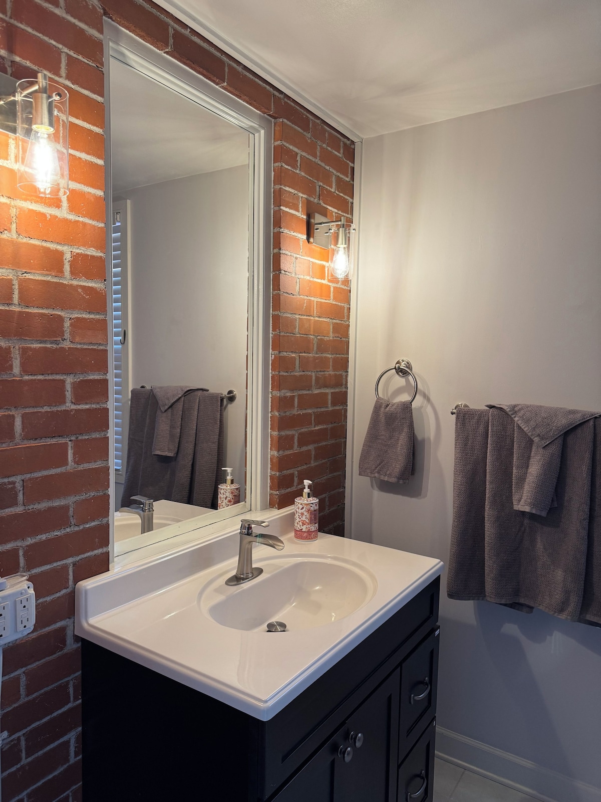A bathroom with a white sink, black cabinets, a large mirror, and a brick wall.