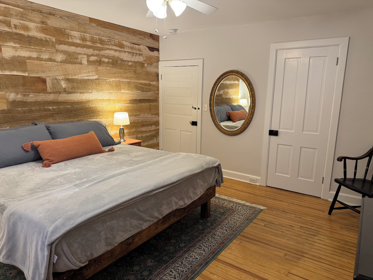 A bedroom with a wooden headboard, a bed with grey bedding, a nightstand with a lamp, a mirror, and a chair.