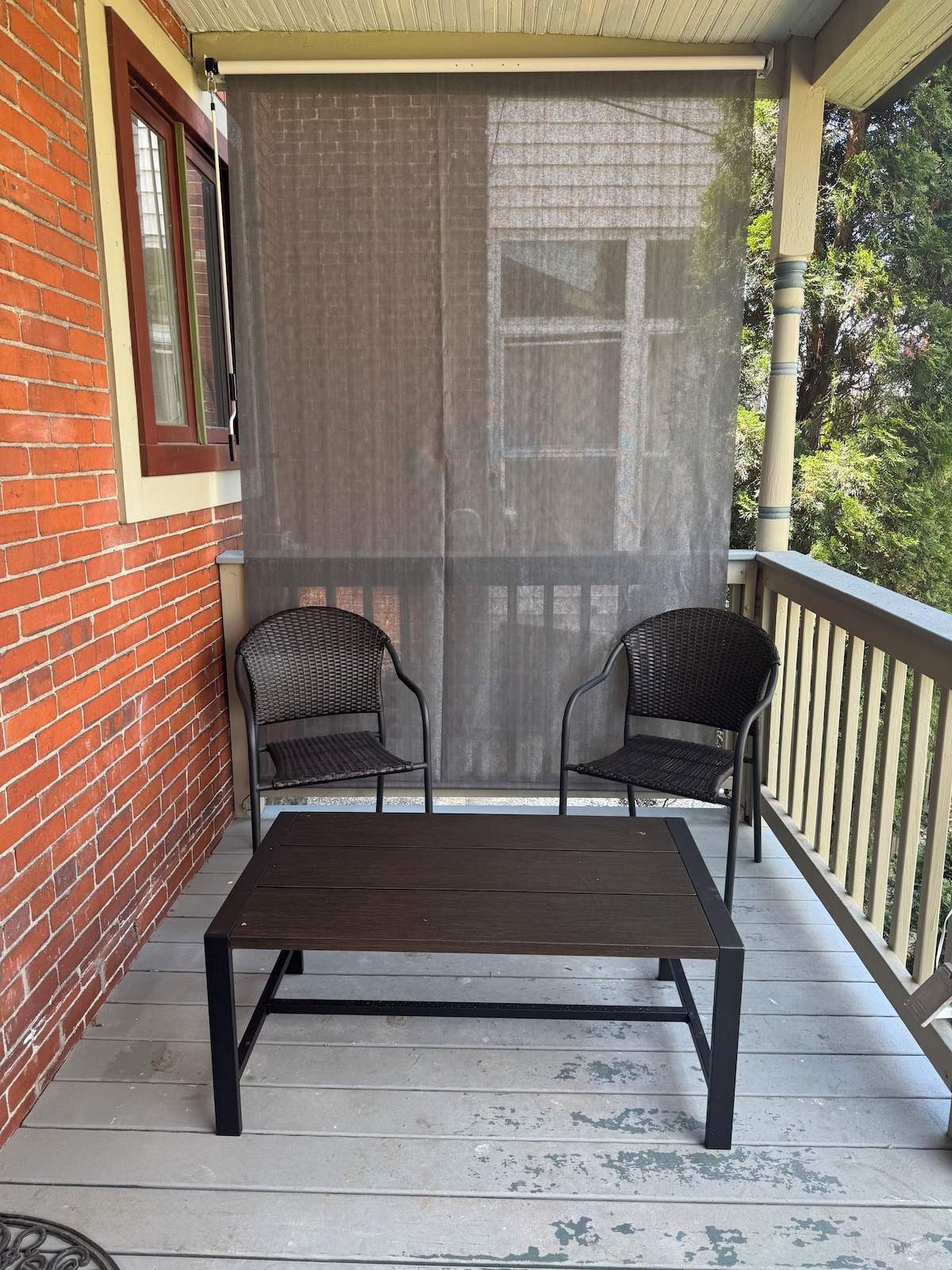A covered outdoor seating area with a table and two chairs, a brick wall, and a window with a curtain.