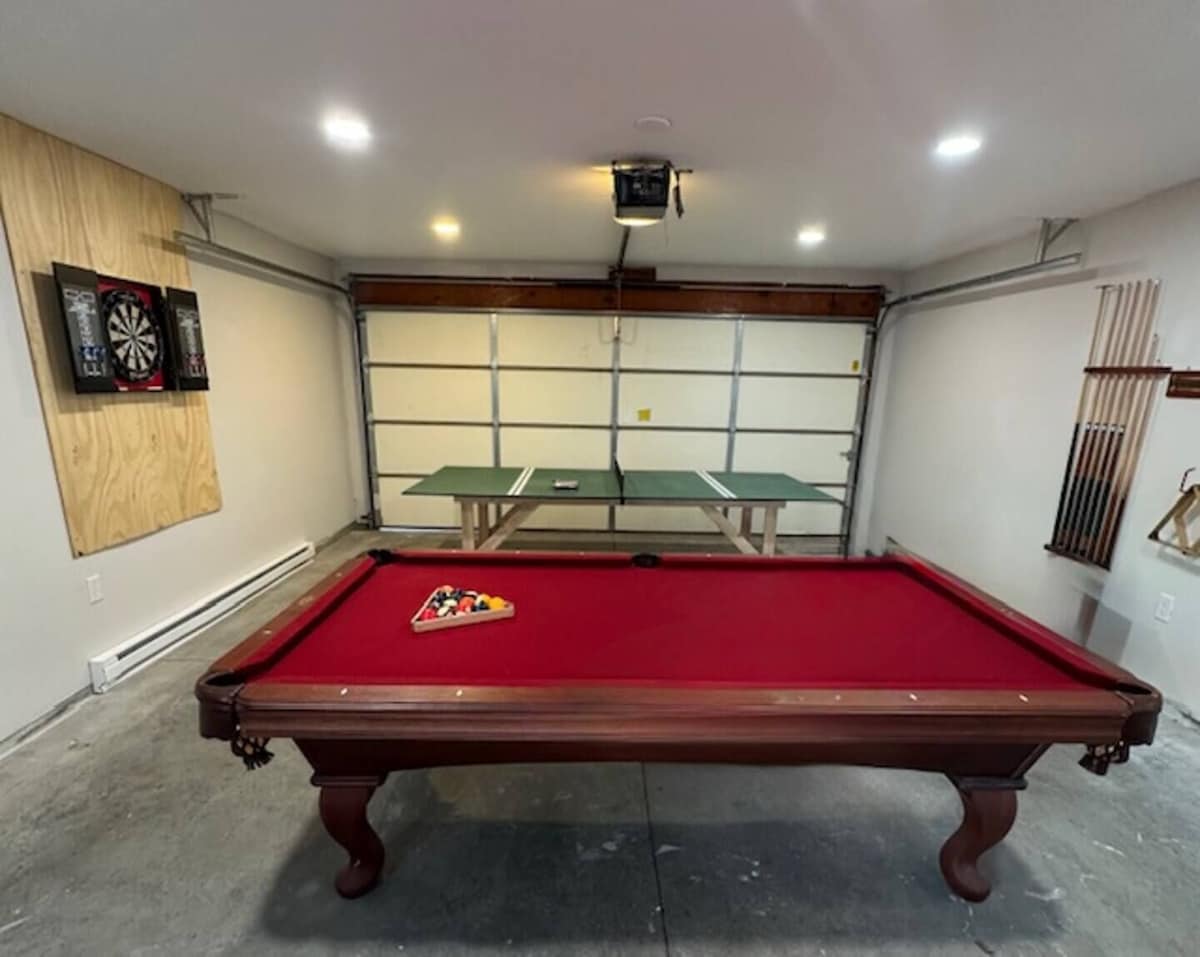 A pool table with a red felt surface, two dartboards on the wall, and a ping pong table in the background.