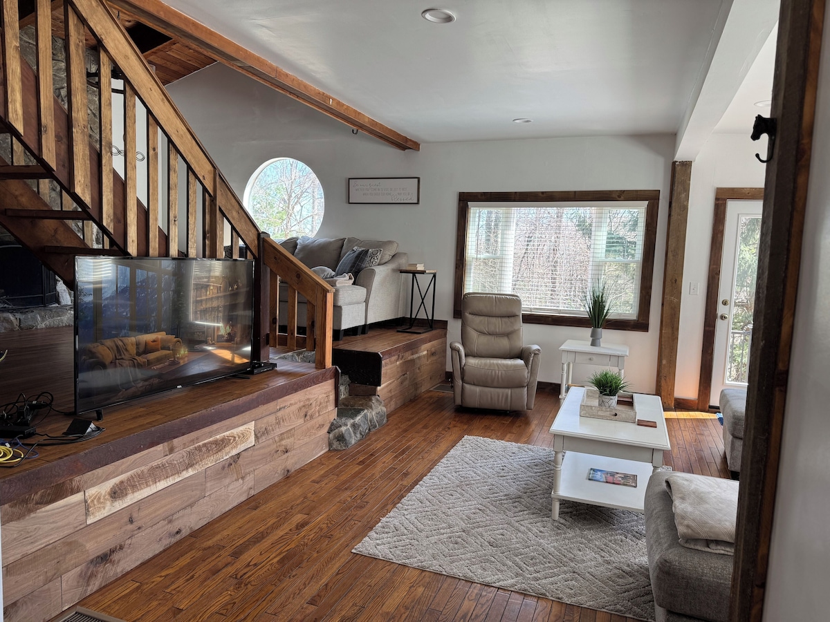 A modern living room with a wooden staircase, a fireplace, a large TV, a comfortable sofa, and a large window with a view of trees.