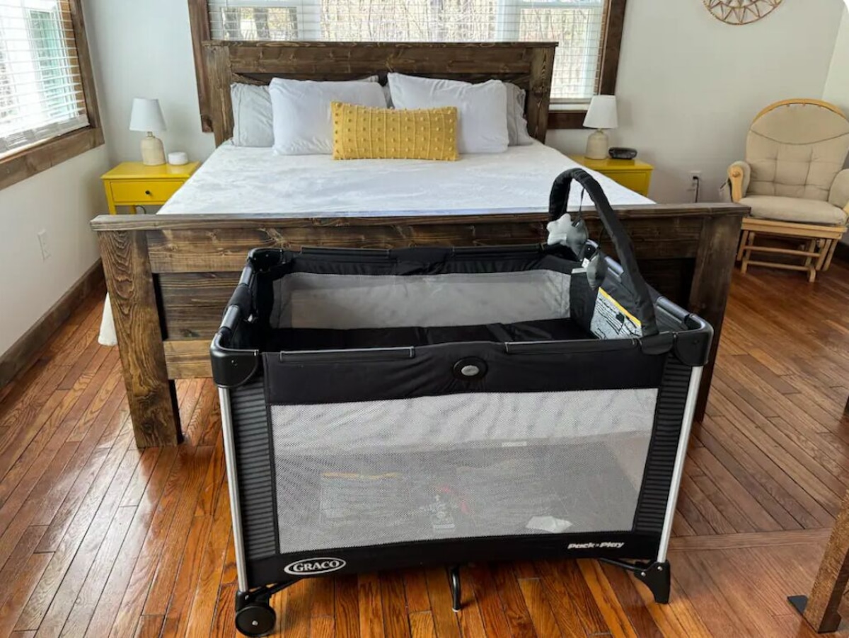 A room with a wooden bed, a small bedside table with a lamp, a chair, and a portable crib.