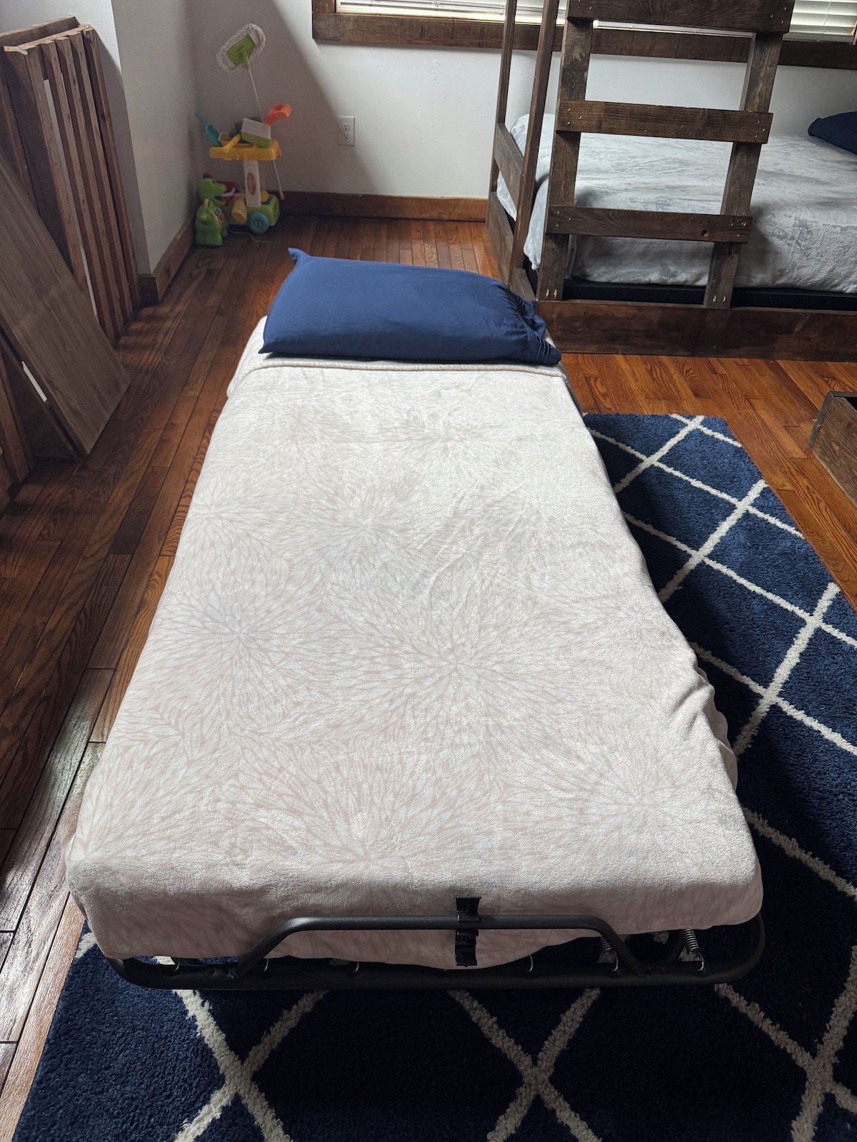 A small, single-bed room with a wooden floor, a blue and white patterned rug, and a window with blinds.