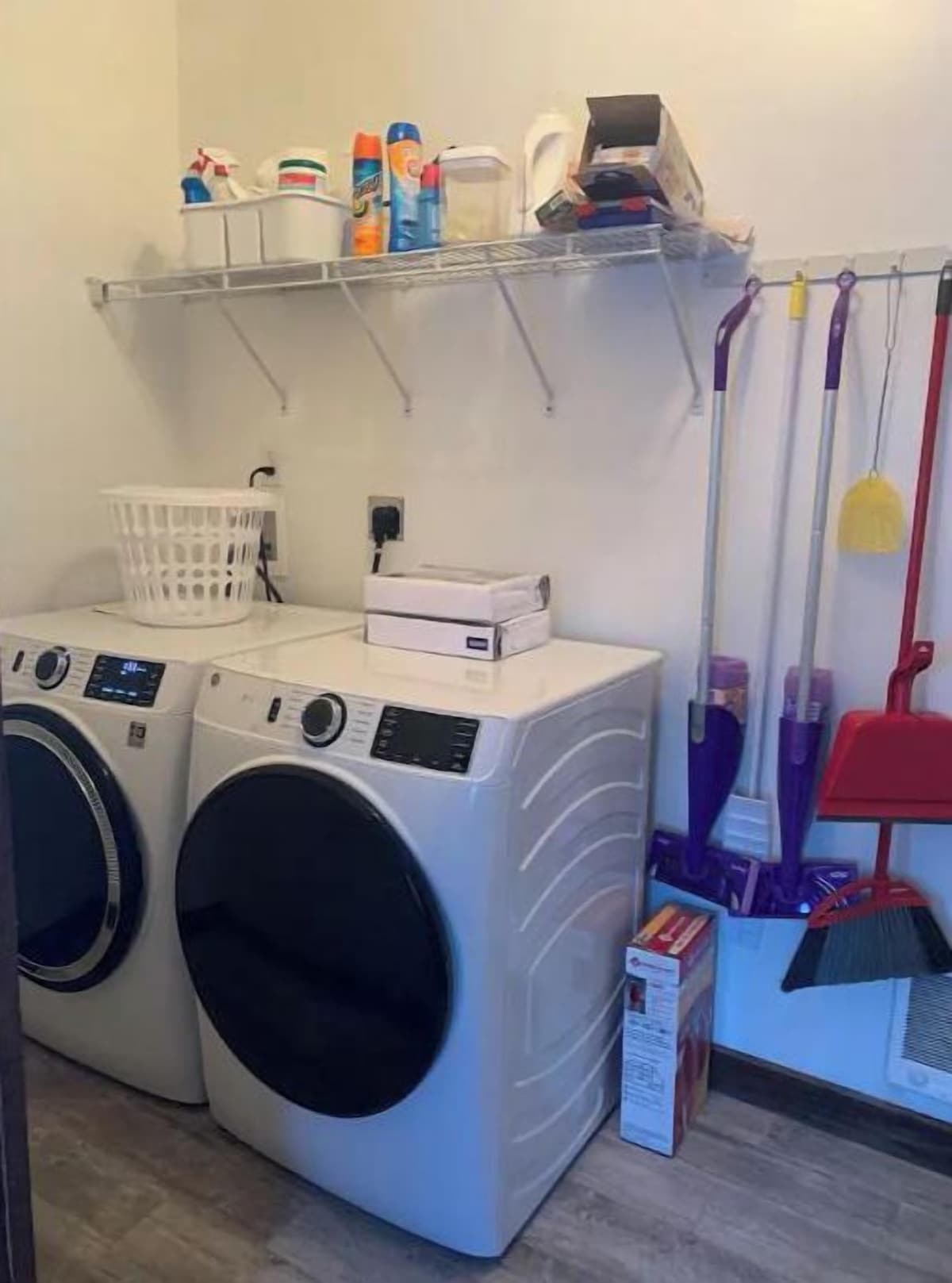 A laundry area with two front-loading washing machines, a drying rack, a shelf with cleaning supplies, and a broom.