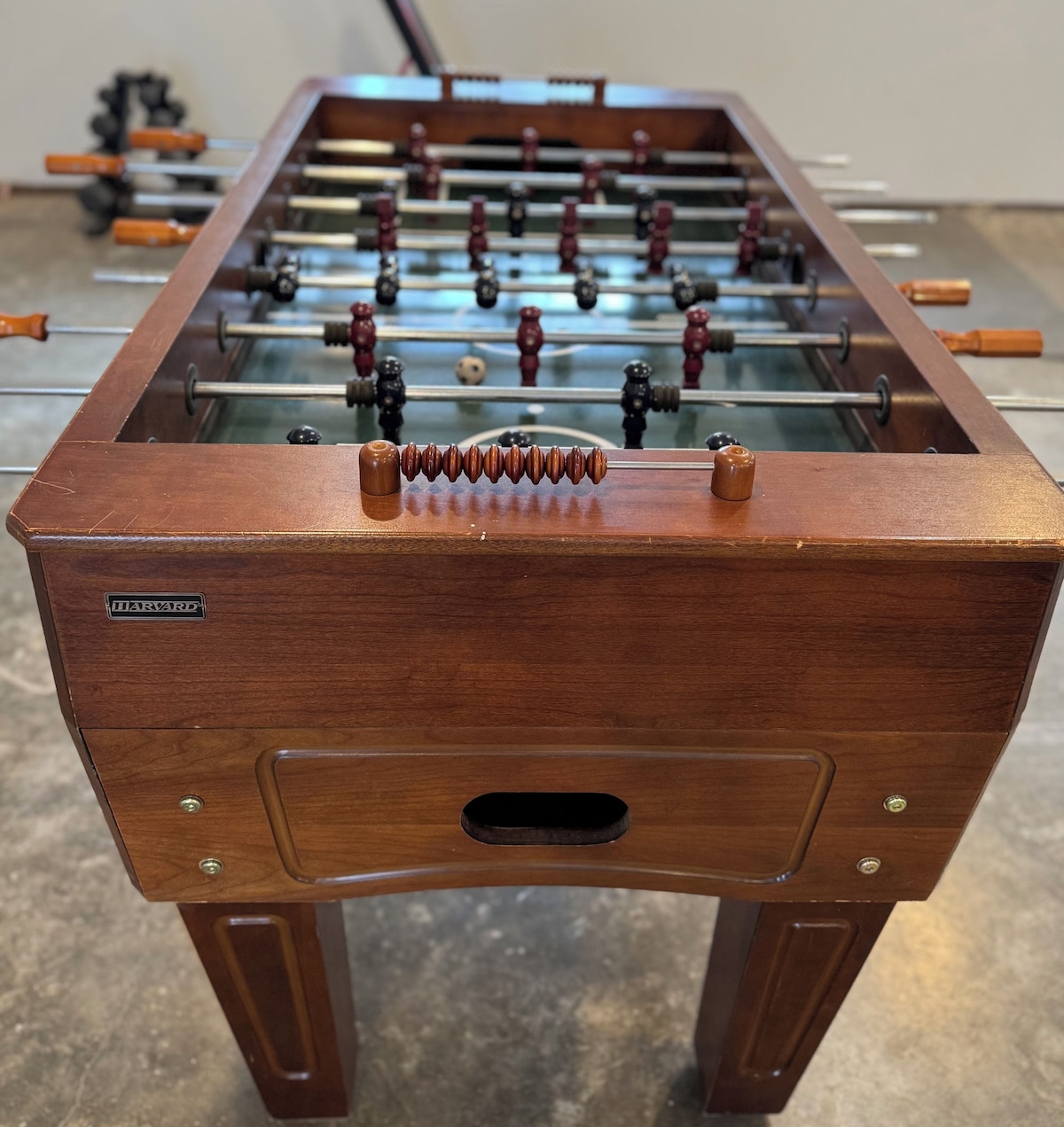 A wooden foosball table with a Harvard logo, featuring a glass top and metal rods with red and black figures.