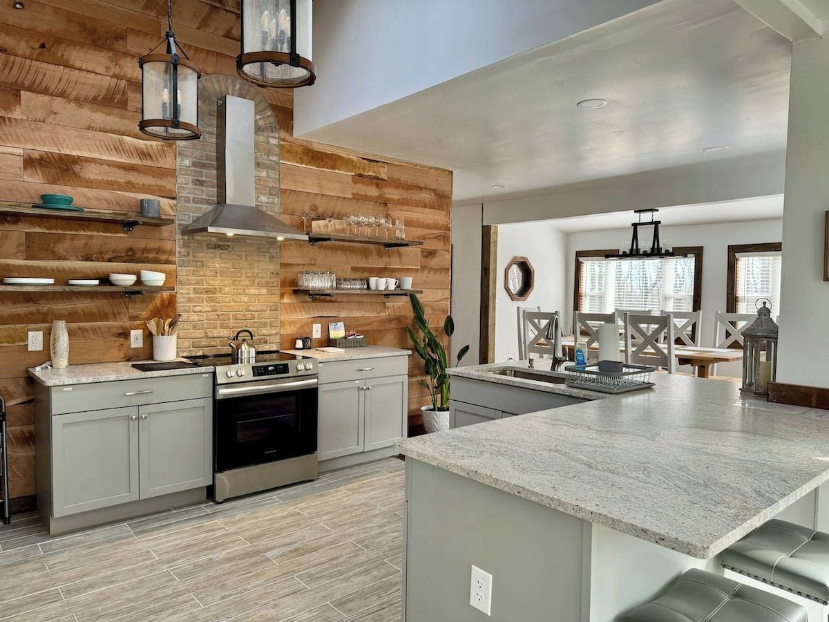 A modern kitchen with a central island, built-in wooden shelving, and a large window providing natural light.