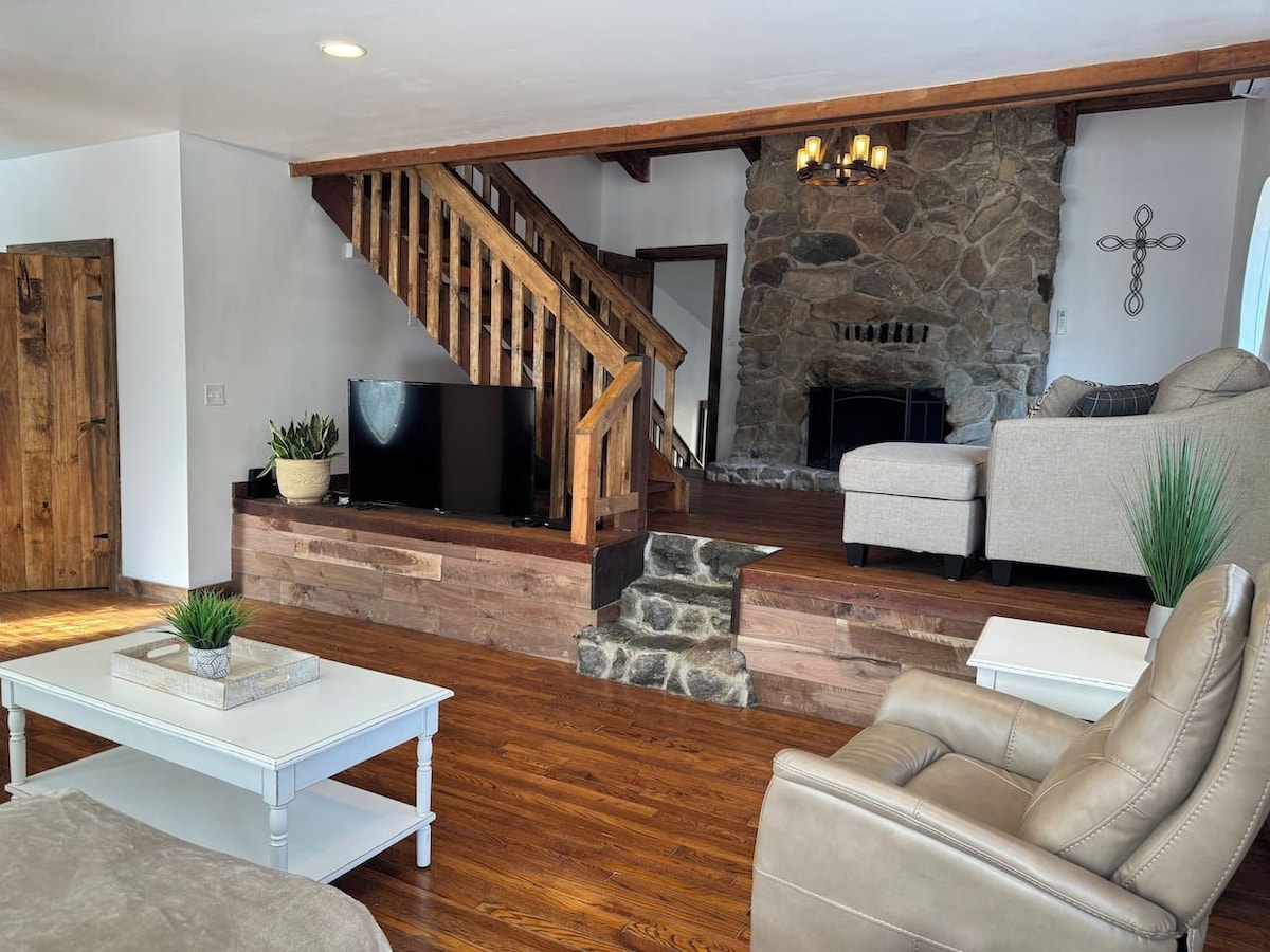 A living room with a stone fireplace, a wooden staircase, a flat-screen TV, and leather furniture.